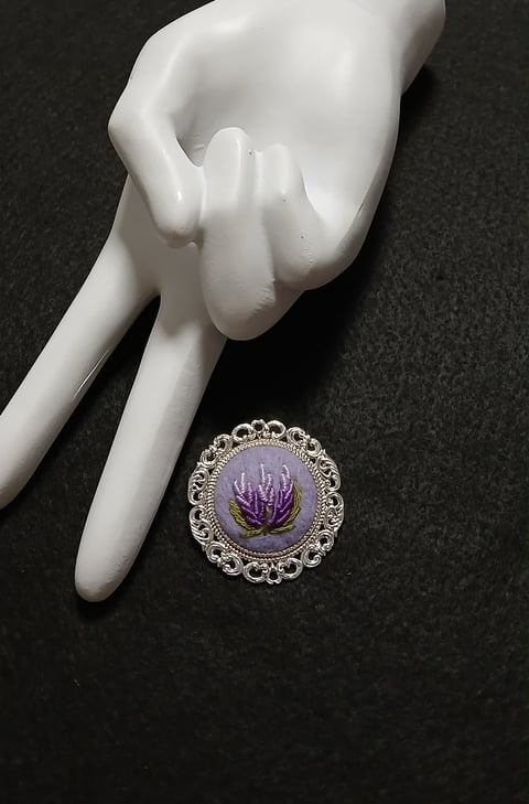 White porcelain hand figurine next to ornate silver brooch with purple dried flower on dark surface