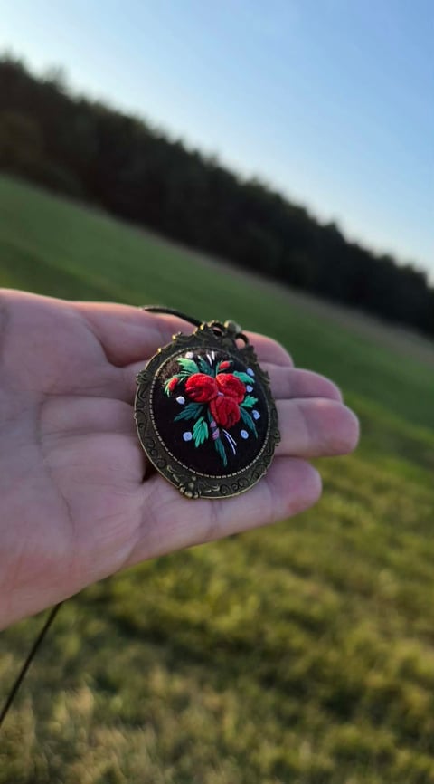 Hand holding ornate embroidered oval pendant with red rose and blue flowers on black fabric, outdoors with green field background