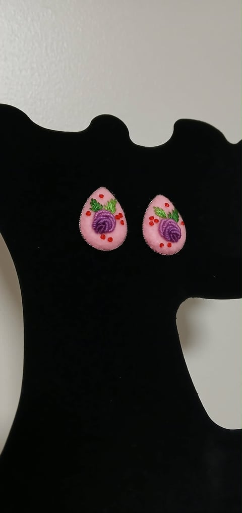 Pair of pink teardrop-shaped earrings with purple flower centers and red berries displayed on black velvet form