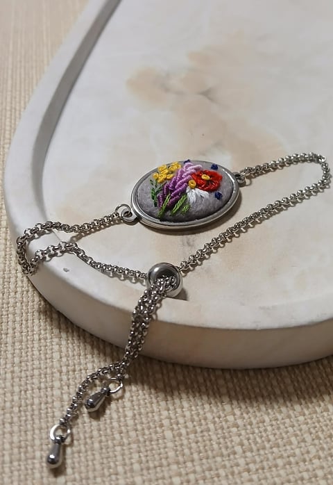 Embroidered floral bracelet with colorful yellow, purple, and red flowers on silver chain resting on white ceramic dish