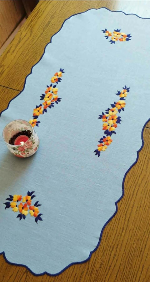 Light blue cloth with scalloped edges and hand-embroidered yellow and purple flower clusters in various arrangements, placed on wooden floor