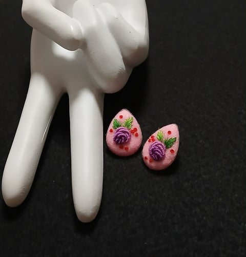 White ceramic hand figurine holding two pink teardrop-shaped floral earrings with purple roses and green leaves on dark background