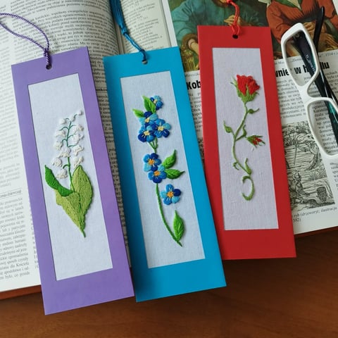 Three colorful bookmarks with pressed flower designs in purple, blue, and red frames displayed on open books