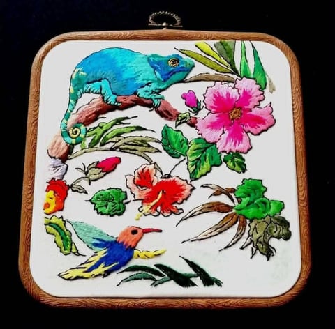 Wooden-framed embroidery artwork featuring a blue chameleon, colorful hummingbird, flowers, and foliage on white fabric