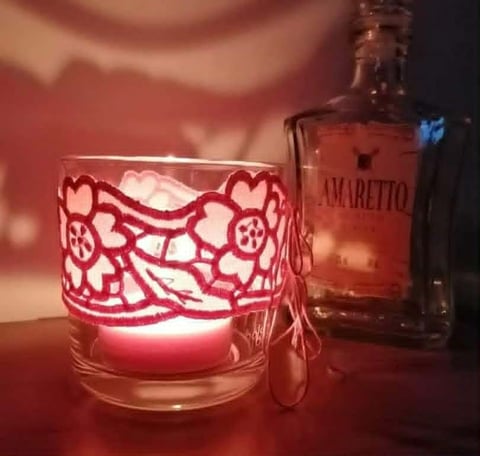 Glowing red candle in decorative glass with floral pattern next to Amaretto bottle on warm background