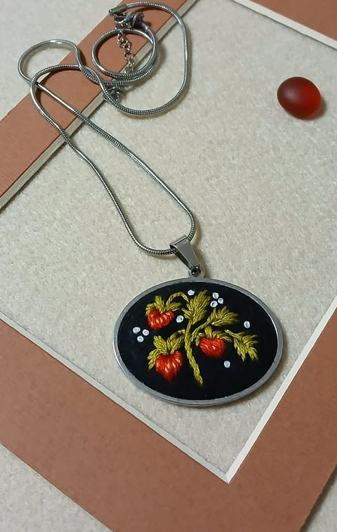 Silver snake chain necklace with oval pendant featuring embroidered strawberries and leaves on black background, displayed with matching rings and red stone