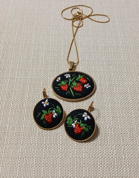 Three embroidered pendants with strawberry and flower designs on black fabric, hanging from gold string on light textured background