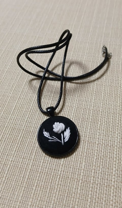 Black pendant necklace with white floral design on circular charm, displayed on beige textured background