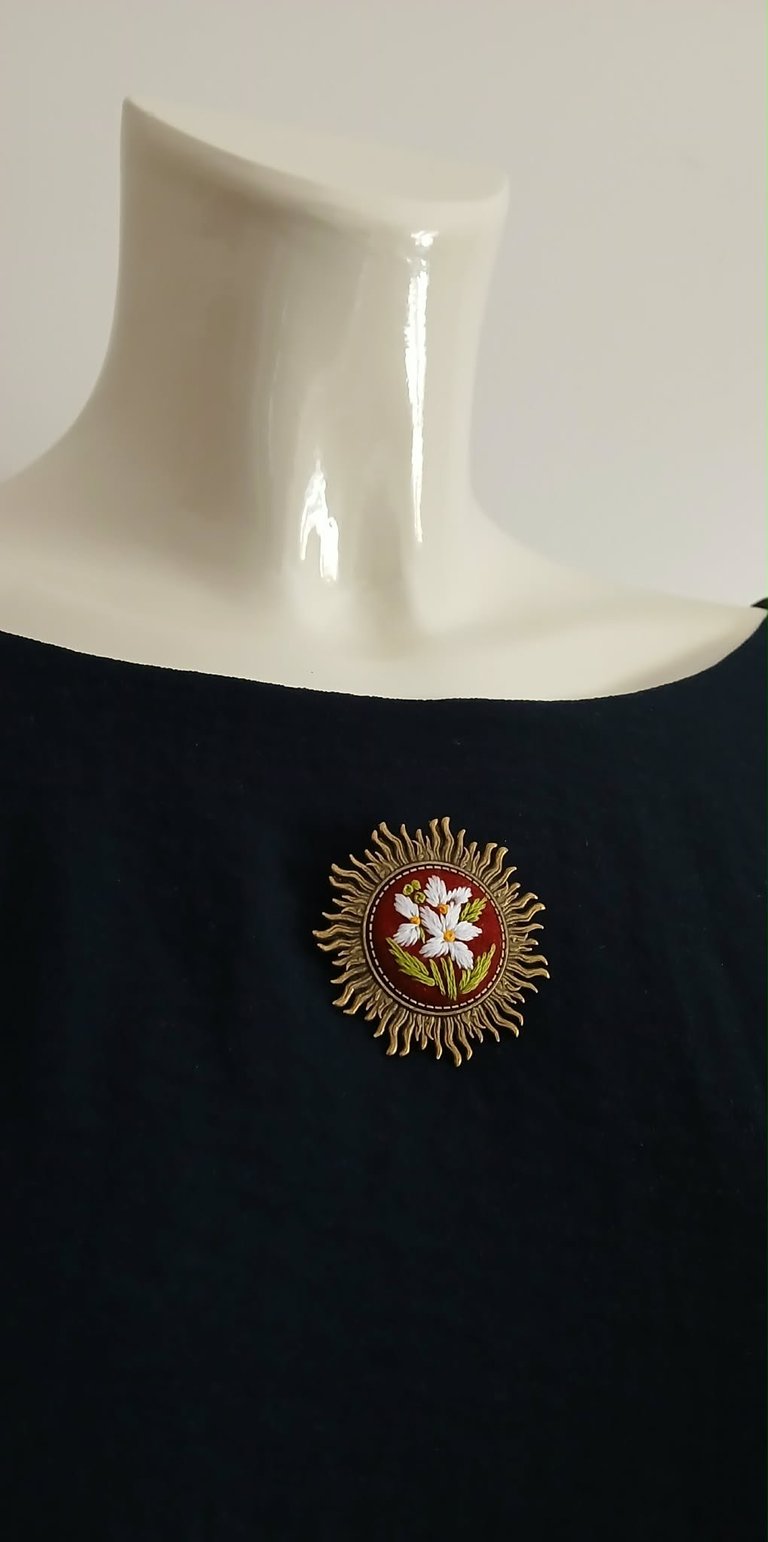 Black sleeveless garment with cream neck collar, displaying embroidered circular floral motif with gold sunburst border and pink flower center