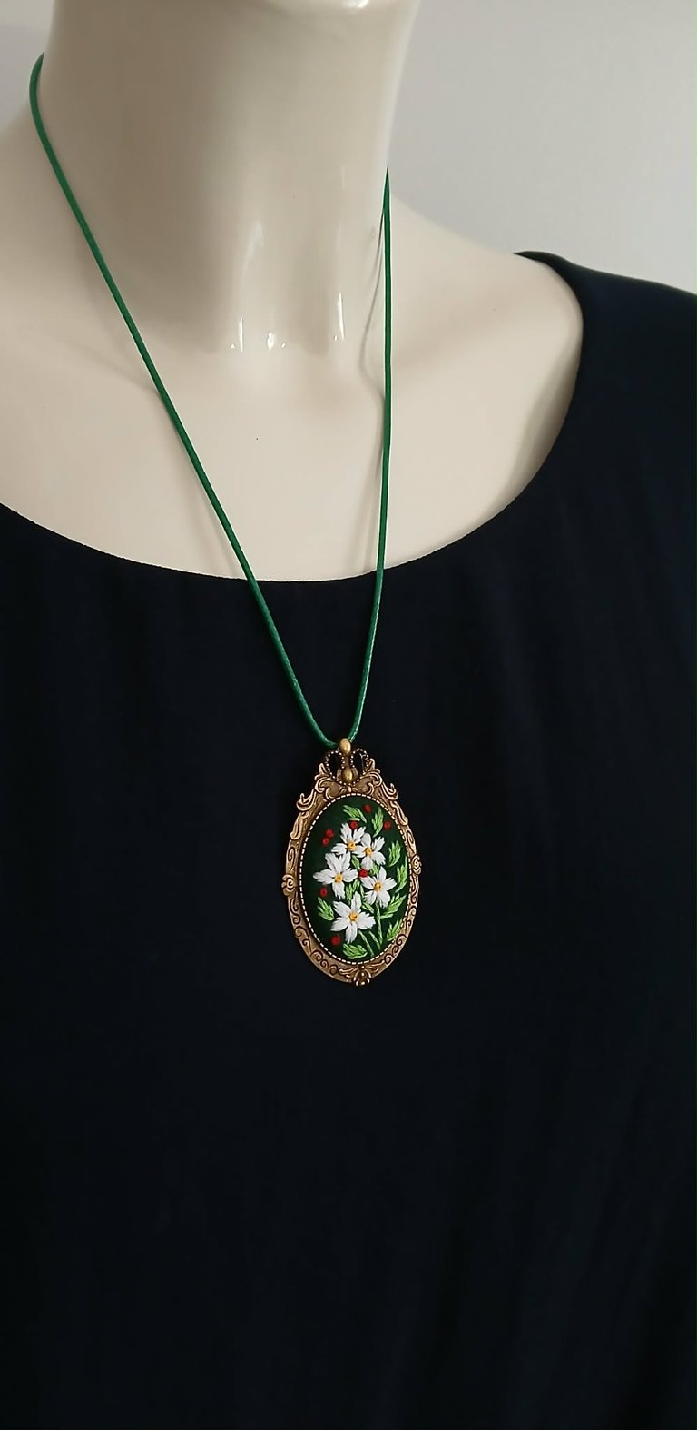 Ornate gold oval pendant with white flowers on green cord necklace worn on mannequin with black dress