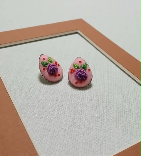 Pair of pink round earrings with purple flowers and green leaves displayed on white fabric in a tan frame
