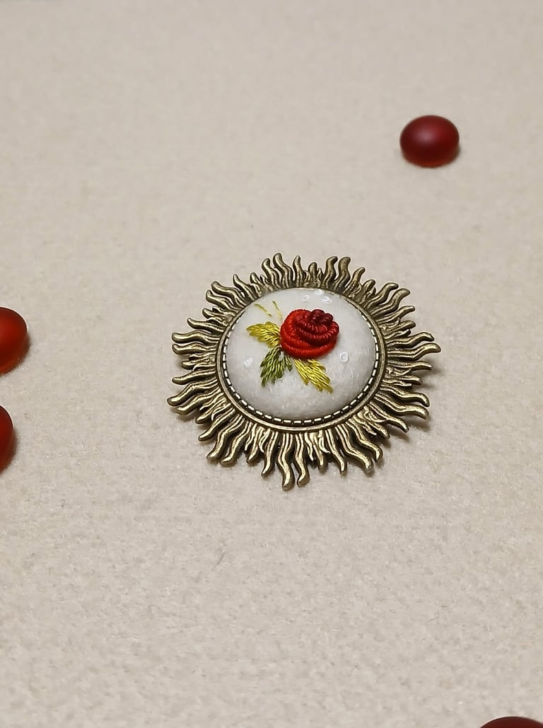Ornate vintage brooch with a red rose illustration on white porcelain surrounded by a decorative brass sun-burst frame, displayed with red beads on a beige background
