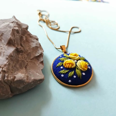 Gold chain necklace with round blue pendant featuring embroidered yellow flowers and pearl details, displayed with a brown stone on light blue background