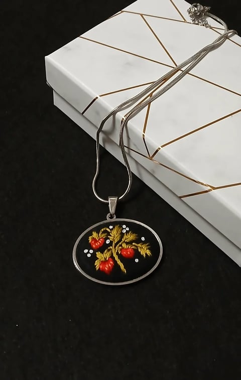 Silver pendant necklace with oval frame containing red tomatoes and yellow leaves on black background, hanging from wrapped white box