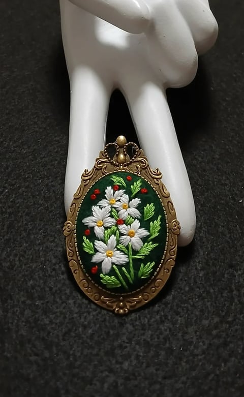 Ornate gold oval pendant with painted white daisies and green leaves on dark background, held in white ceramic hand