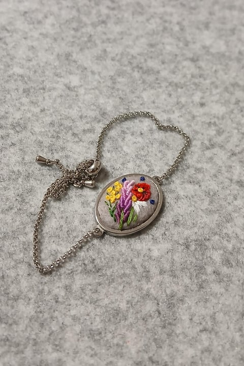 Silver chain necklace with round pendant featuring embroidered yellow, purple, and red flowers on white fabric background