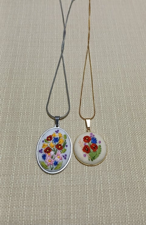 Two embroidered flower pendant necklaces with gray and gold chains on a light textured background.