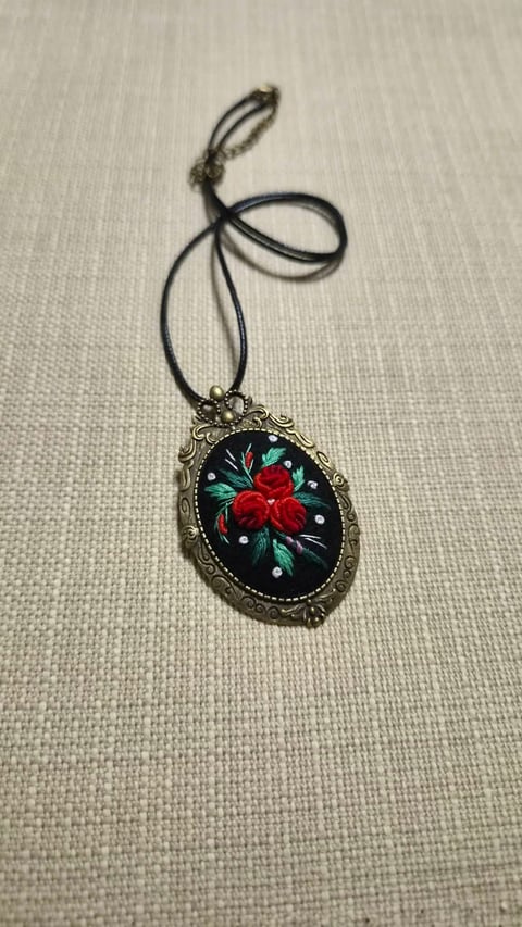 Ornate pendant necklace with red rose design on black background, hung by black cord on beige fabric