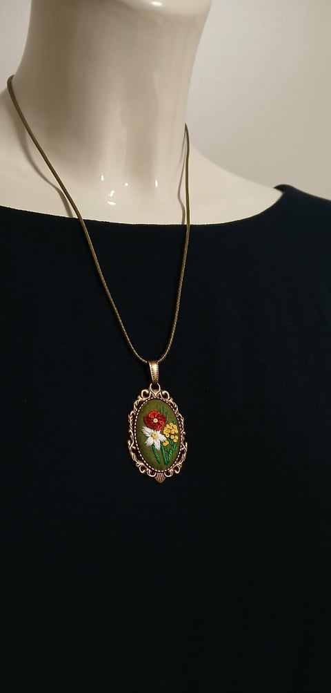 Gold necklace with ornate oval pendant featuring pressed flowers in red, white, and green, worn on mannequin with black top