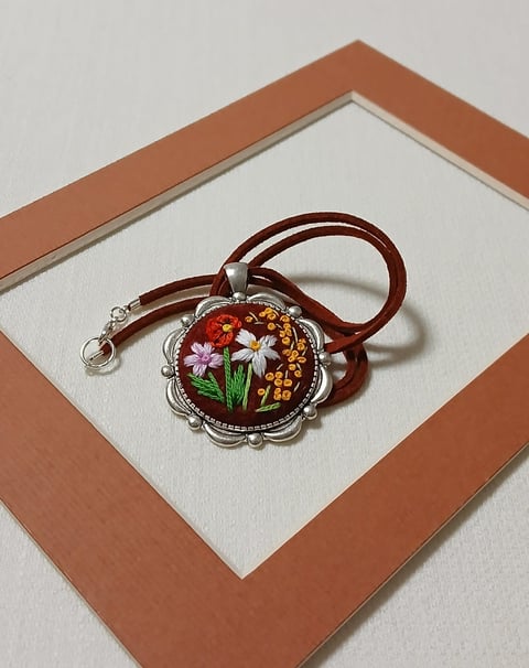 Decorative pendant necklace with floral embroidery design on brown cord, displayed on cream and rust-colored matting