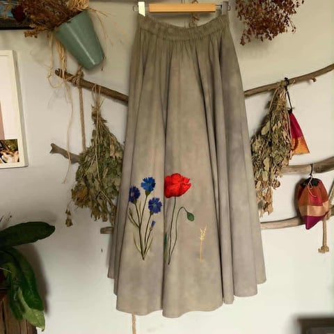 Gray linen skirt with embroidered colorful flowers hanging on wooden rod against wall with dried plants and botanical decor