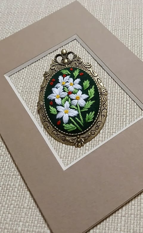 Hand-painted ornate brooch featuring white daisies and green leaves on black oval pendant in decorative bronze frame
