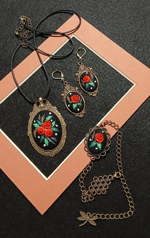 Vintage-style jewelry set featuring red rose designs on black enamel with gold accents, including a necklace, earrings, and bracelet displayed on a peach and black mat.