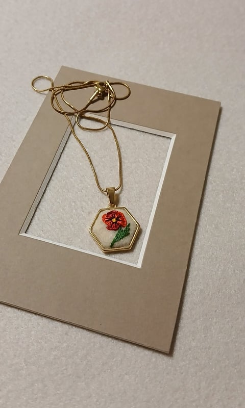 Gold chain necklace with hexagonal pendant featuring embroidered red poppy flower displayed on tan cardstock with white mat
