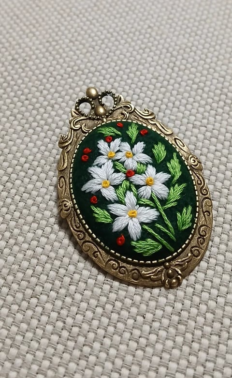 Ornate oval pendant with white daisies, red berries, and green leaves embroidered on dark background, featuring decorative bronze frame and pearl bow top