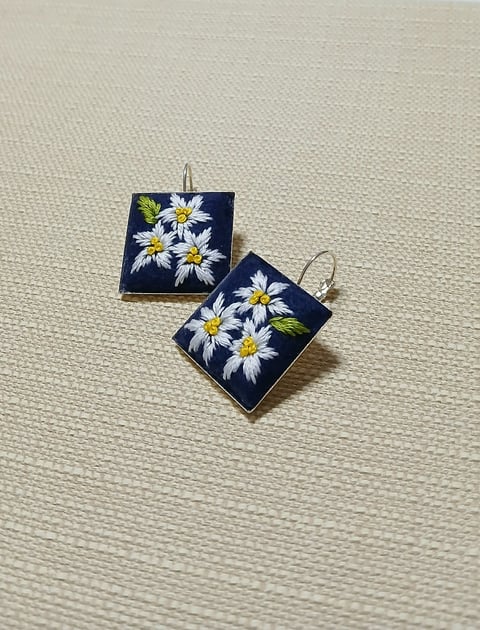 Pair of square earrings with white daisies and yellow centers on dark blue background