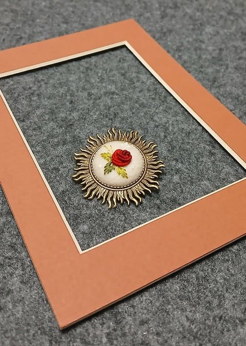 Ornate vintage brooch with red rose design and gold spiked backing, displayed in coral mat frame on gray felt