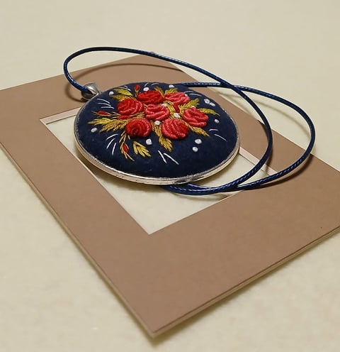 Embroidered round pendant with red roses on navy blue fabric, attached to a blue cord necklace on a tan display mat