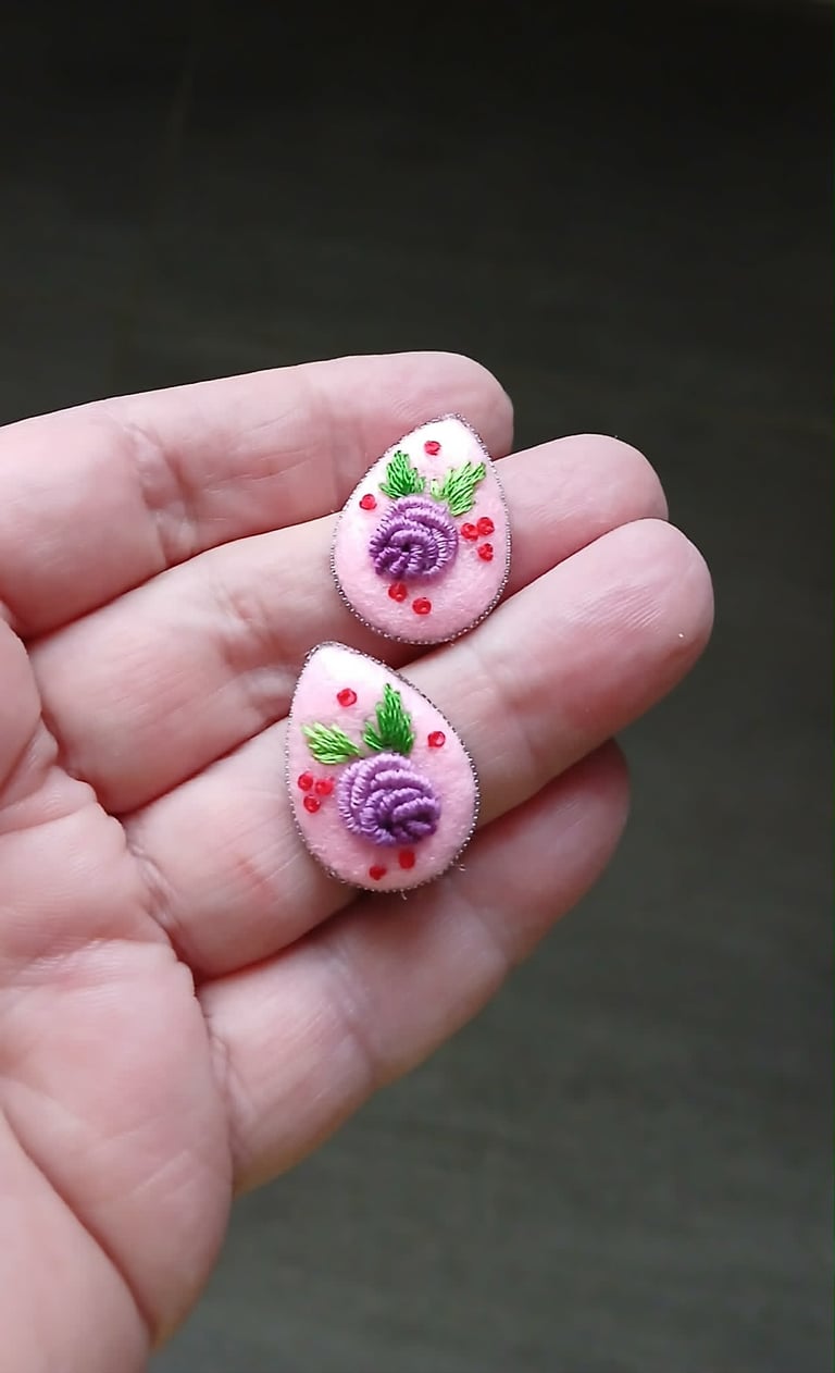 Two embroidered teardrop-shaped brooches held in a hand, featuring purple flowers and red berries with green leaves on pink fabric