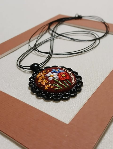 Decorative pendant necklace with embroidered floral design on black scalloped base, displayed on tan cardstock
