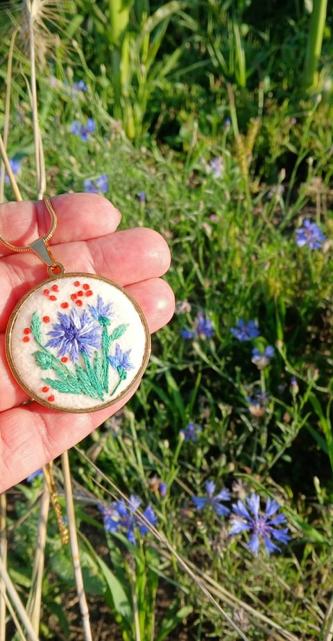 Hand holding embroidered pendant with purple and blue flowers and red berries, displayed against blooming wildflower garden background