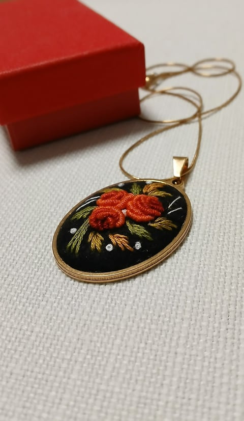 Gold oval pendant necklace with red raspberries and green leaves on black enamel background, displayed with red gift box