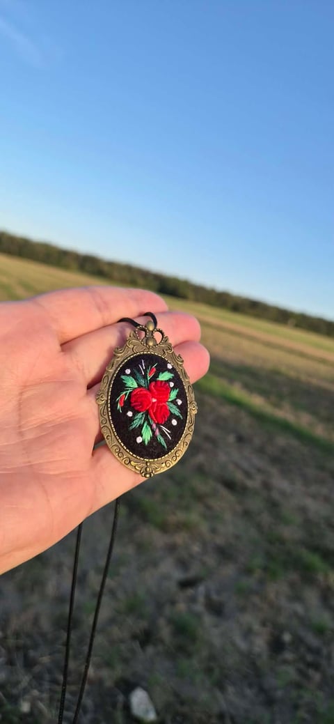 Hand holding ornate oval pendant with embroidered red flower and green leaves on black background in open field