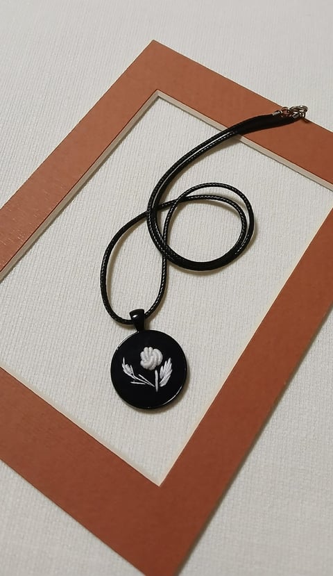 Black pendant necklace with white floral design on cream paper, mounted on rust-colored frame
