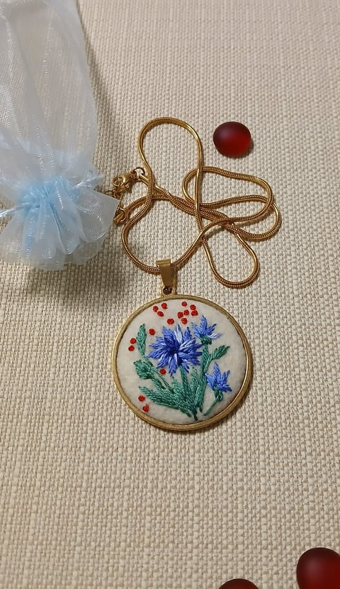 Gold pendant necklace with embroidered floral design featuring blue flowers and red berries on cream background, displayed on beige fabric with decorative beads