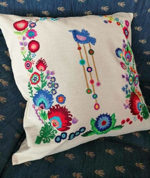 Embroidered cream-colored decorative pillow with colorful floral borders and a blue bird with hanging beads on dark patterned background