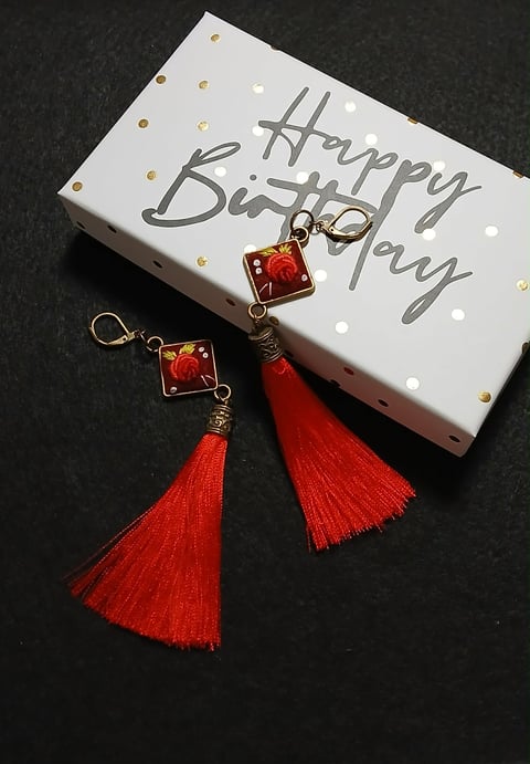 White Happy Birthday box with gold polka dots and pair of red tassel earrings with decorative charms on dark background