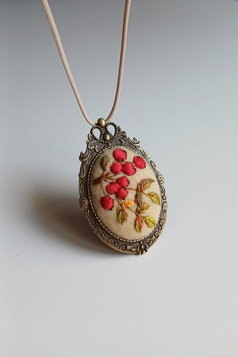 Ornate vintage pendant necklace with oval bronze frame containing embroidered red berries and tan leaves on cream fabric, hanging from beige cord.