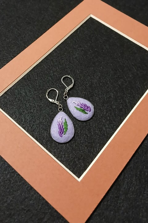 Pair of lavender teardrop earrings with purple flower design and silver hooks on display against peach and black background