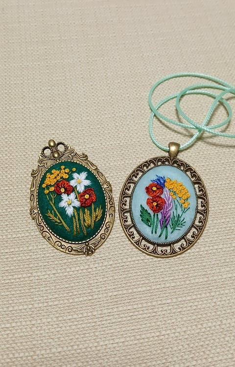 Two embroidered floral pendants with ornate bronze frames on a teal cord, displaying colorful wildflowers on cream and green backgrounds.