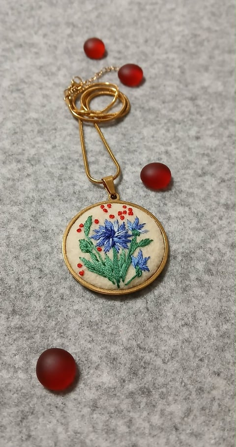 Gold pendant necklace with embroidered circular charm showing blue flowers and red berries, surrounded by four dark red beads on gray surface