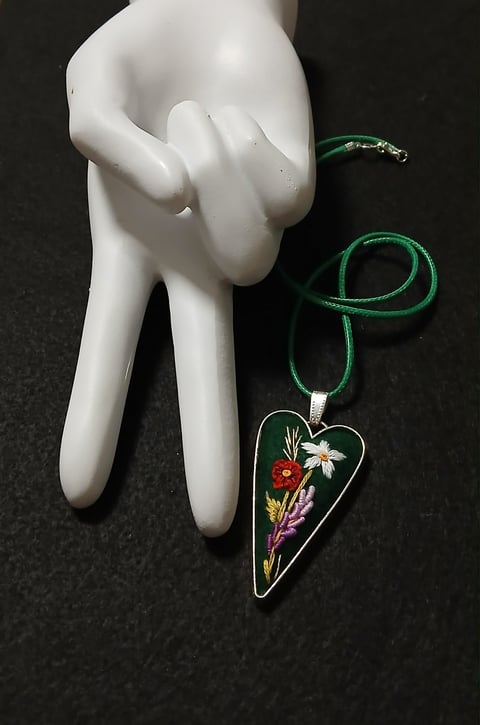 White mannequin hand displaying a green cord necklace with a floral heart pendant featuring embroidered flowers on dark background