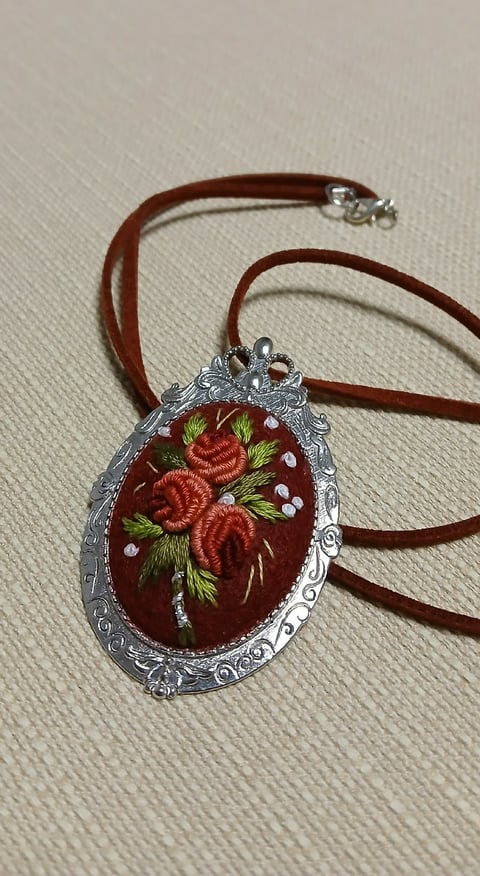 Ornate silver pendant with embroidered red roses on burgundy fabric, hanging from a brown cord
