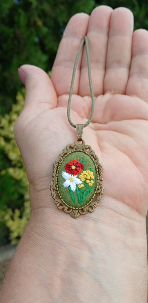 Hand holding a decorative oval pendant necklace with embroidered red, white, and yellow flowers on green background
