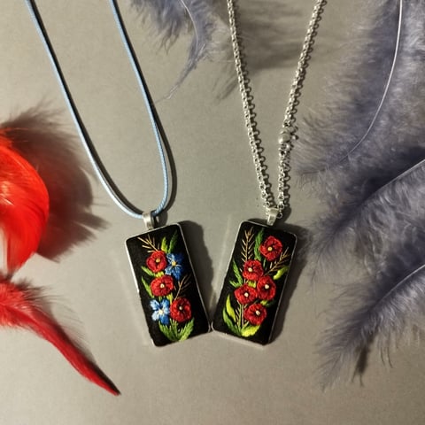 Two decorative pendant necklaces with floral designs on black enamel rectangles, hung on dark and silver chains against a neutral background.