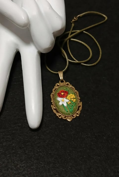 Gold ornate oval pendant with red and yellow embroidered flowers on green background, suspended from gold chain next to white mannequin hand